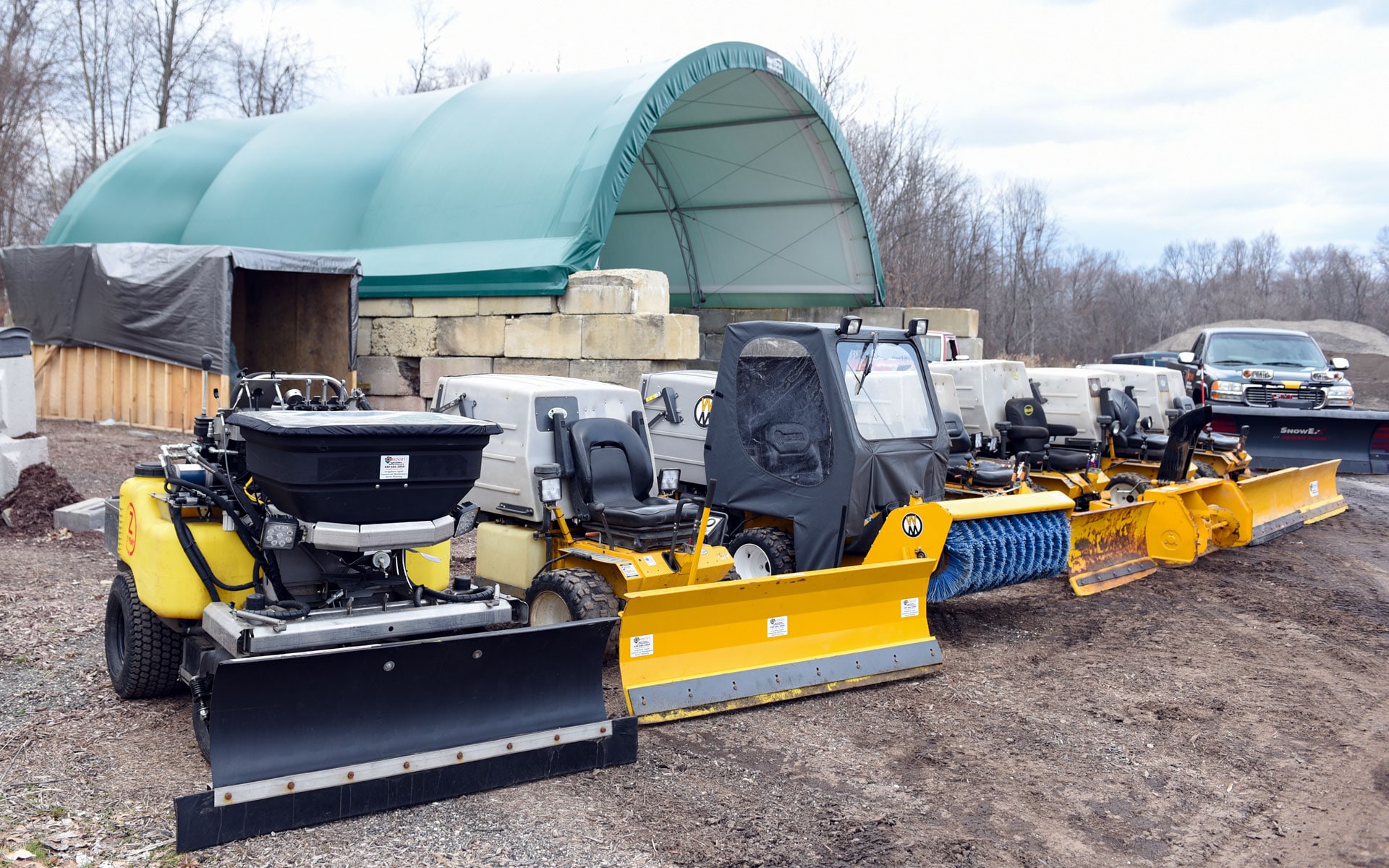 A salt spreader, compact machines with plow blades and a rotary broom staged in the yard